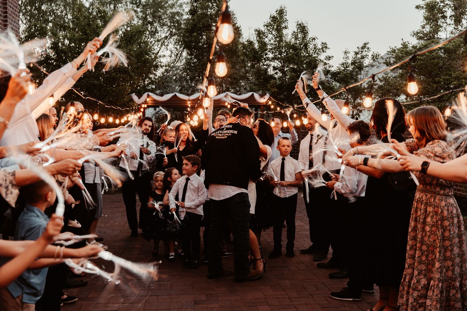 Couple shares romantic kiss during wedding send-off with guests holding streamers under string lights at dusk.