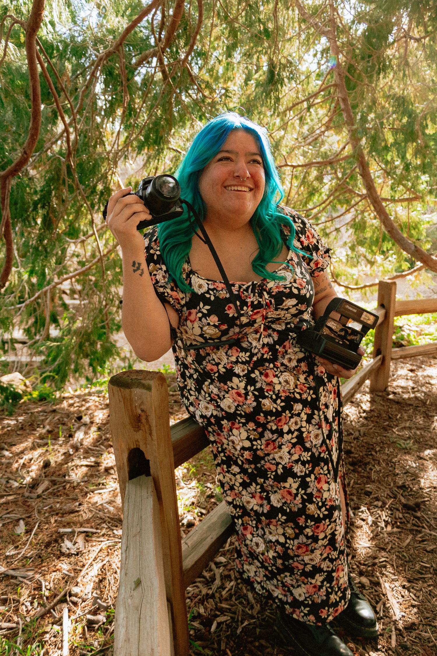 A Mexican woman photographer in a floral dress and teal hair holds vintage cameras while standing by a wooden railing in dappled sunlight.