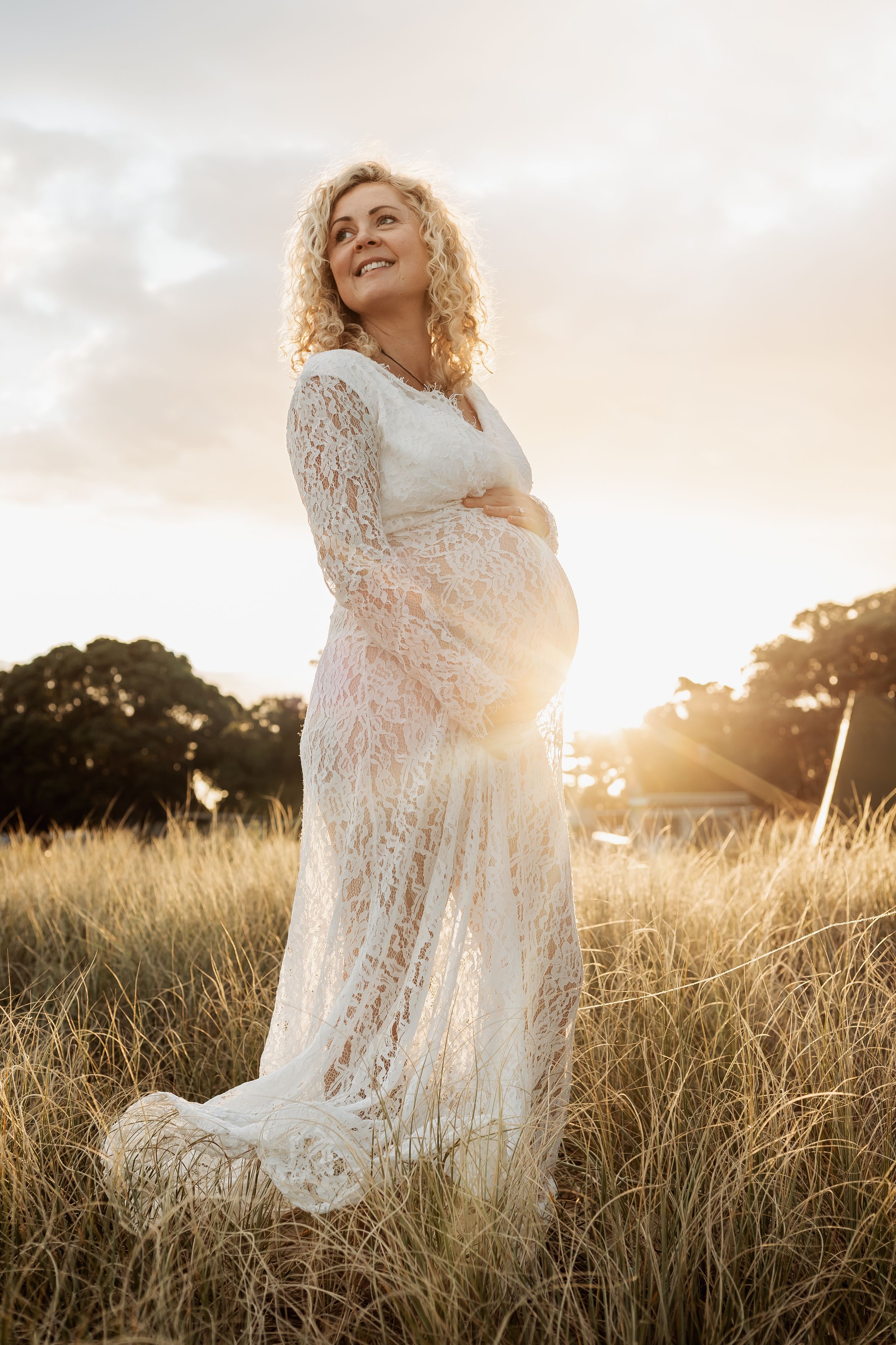 Smiling pregnant woman in white lace dress standing in golden field at sunset.