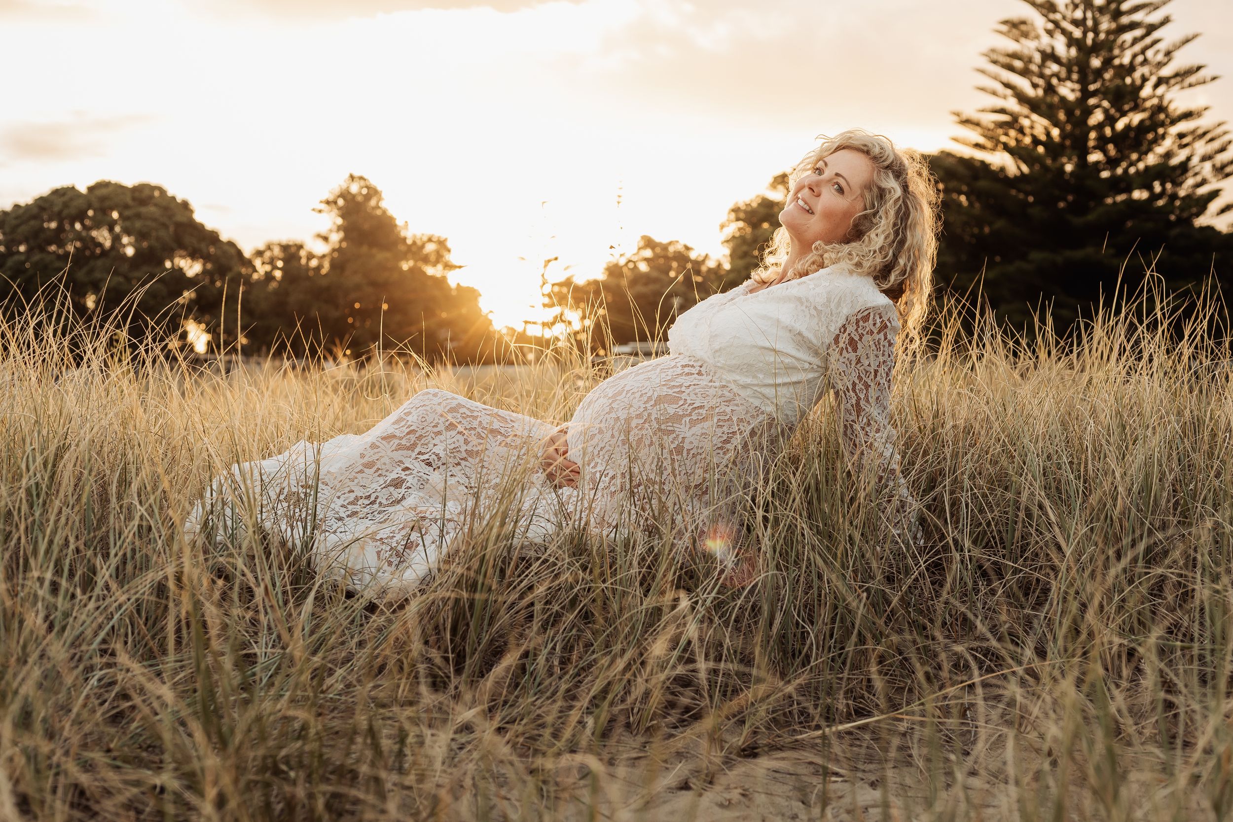 Pregnant woman in white lace dress reclining in golden grass at sunset.