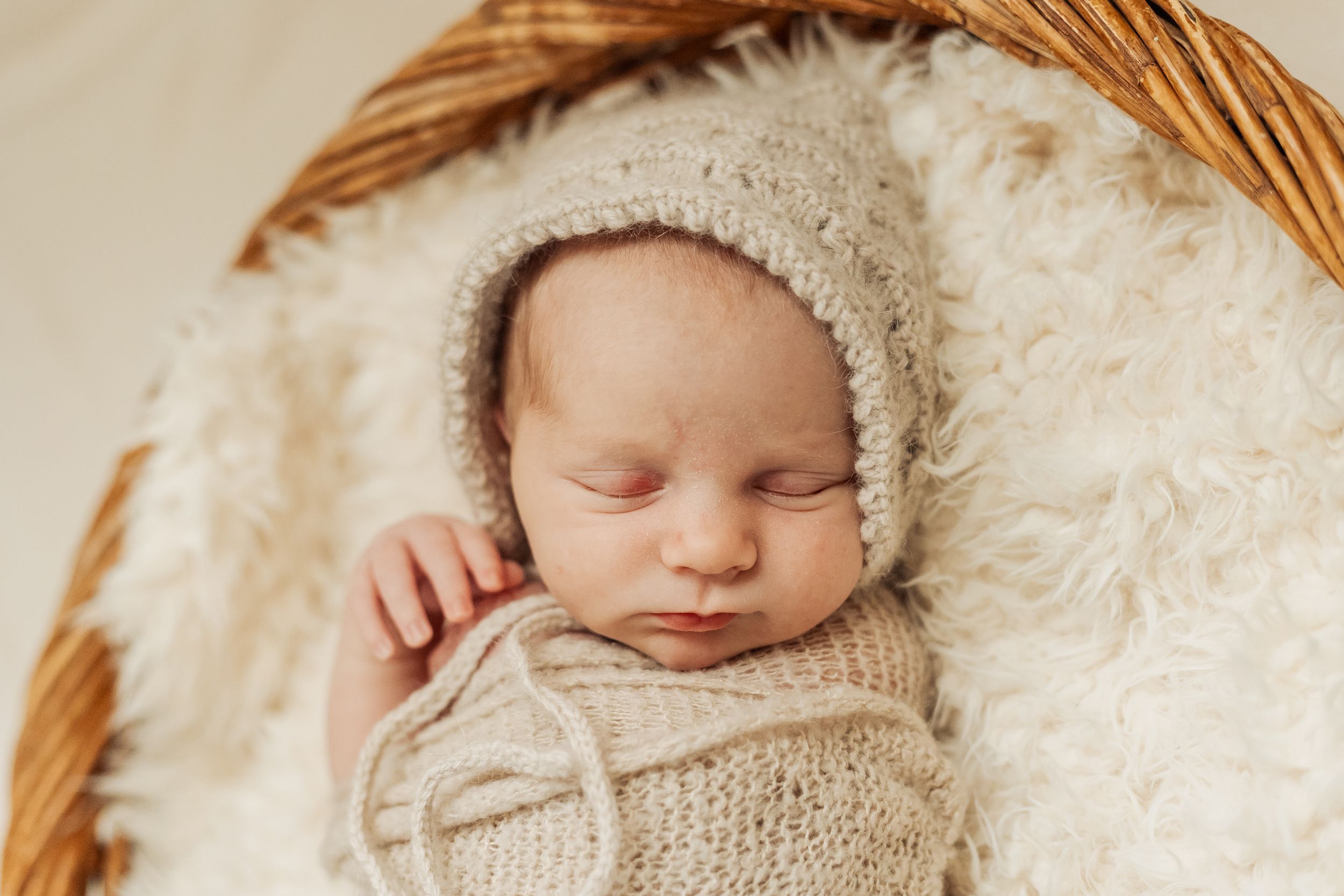 Sleeping newborn baby wrapped in knit blanket and bonnet in a wicker basket.