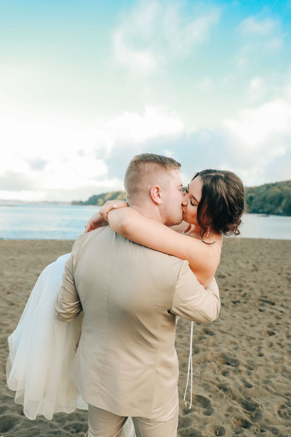 Groom carrying bride in white dress sharing a kiss on a sandy beach.