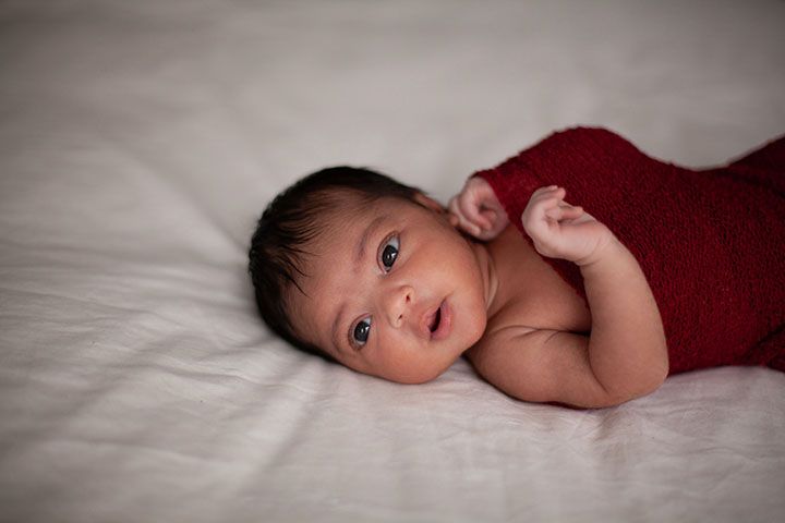 A baby in a red wrap lies on white bedding while looking at the camera.
