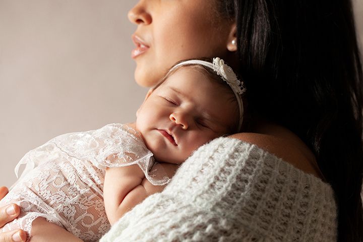 Sleeping newborn in white lace dress rests peacefully against caregiver in knitted sweater.