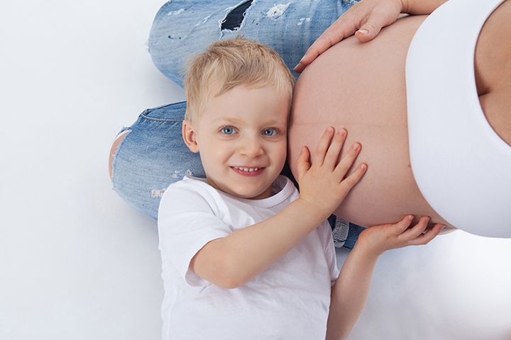Young child in white t-shirt places hands on pregnant belly while laying against white background.