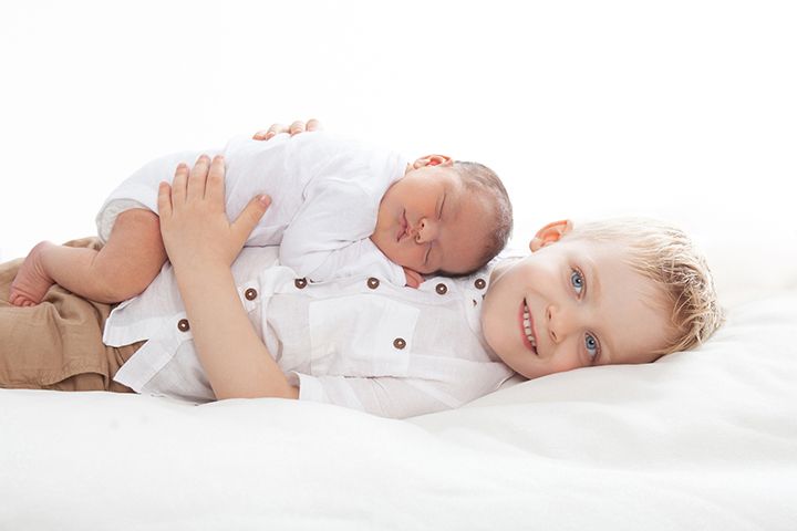Two young siblings in white clothing lie together on a white background with one gently holding the other.