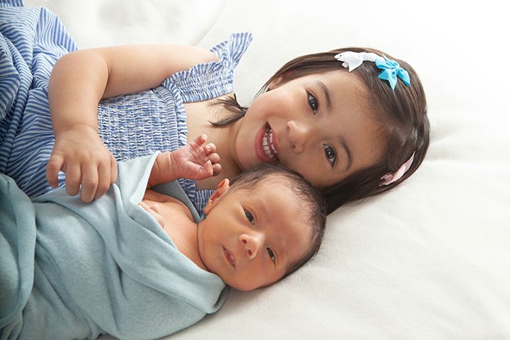 Two young children laying together on white bedding wearing blue clothing and smiling warmly.