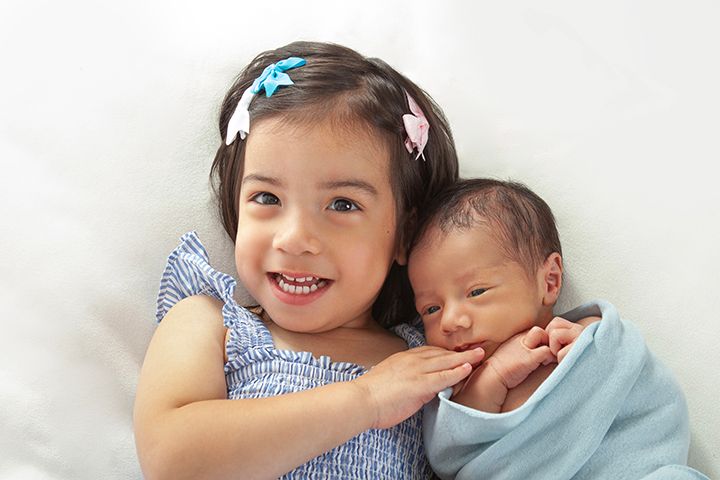 Happy big sister in blue dress gently snuggles with sleeping newborn sibling wrapped in soft blanket.