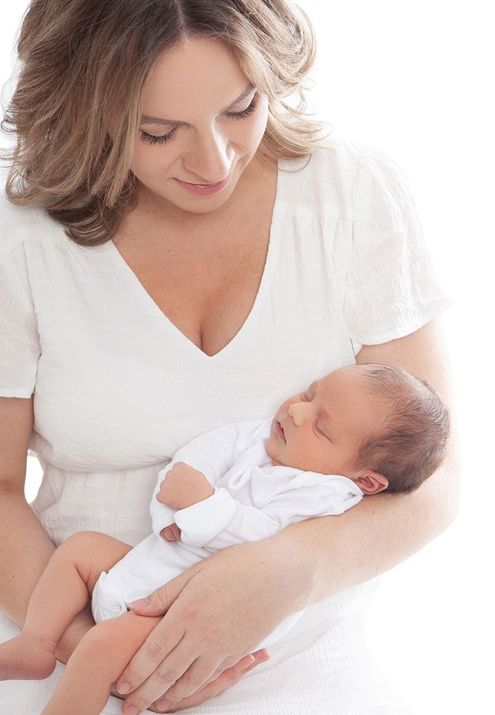 A person in white clothing tenderly holds and gazes at a sleeping newborn baby swaddled in white.