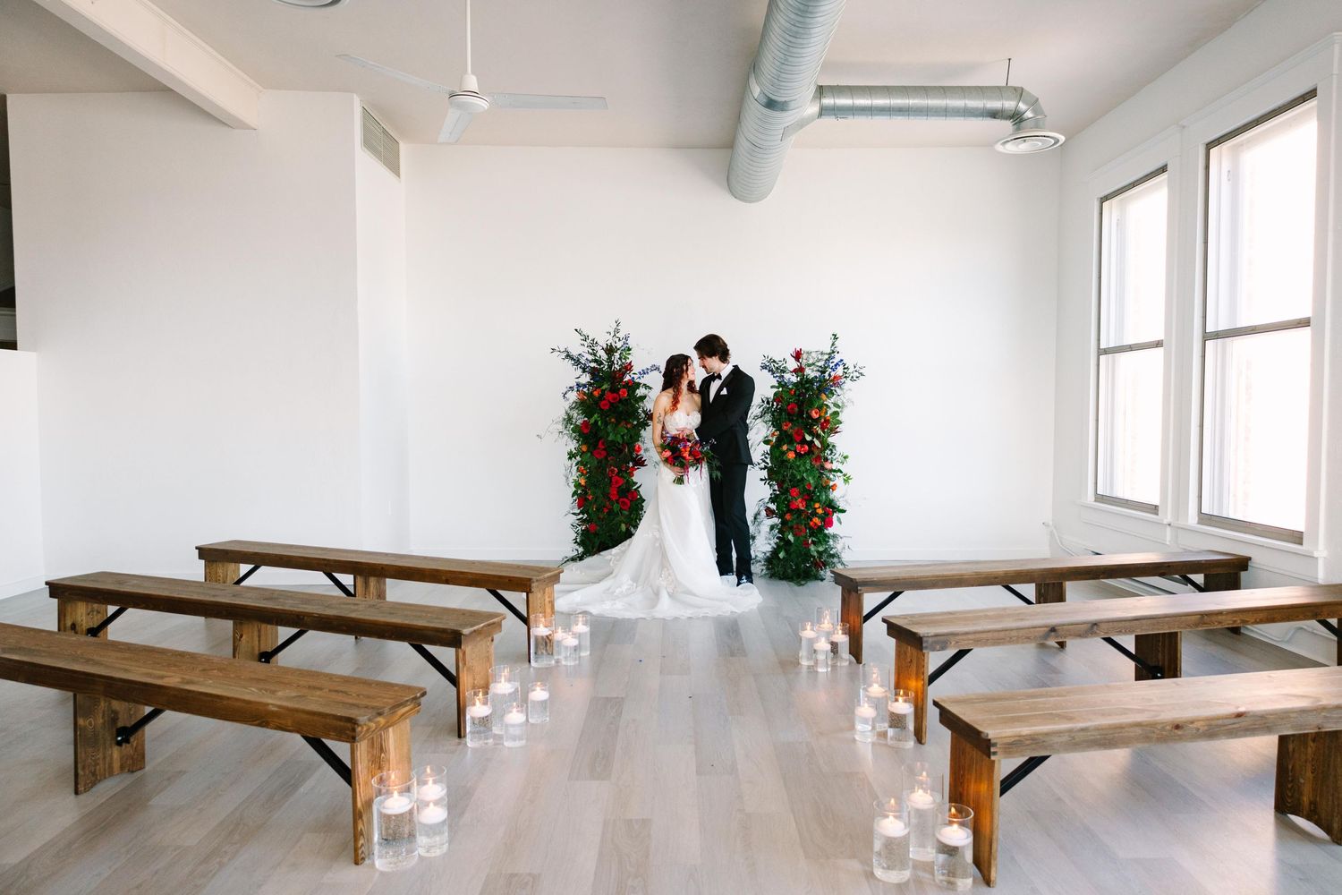 Modern minimalist wedding ceremony setup with wooden benches and greenery arch in bright white interior.