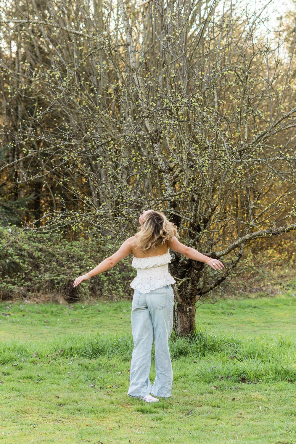 Someone in a white top and light jeans stands with arms outstretched on a grassy lawn near autumn trees.