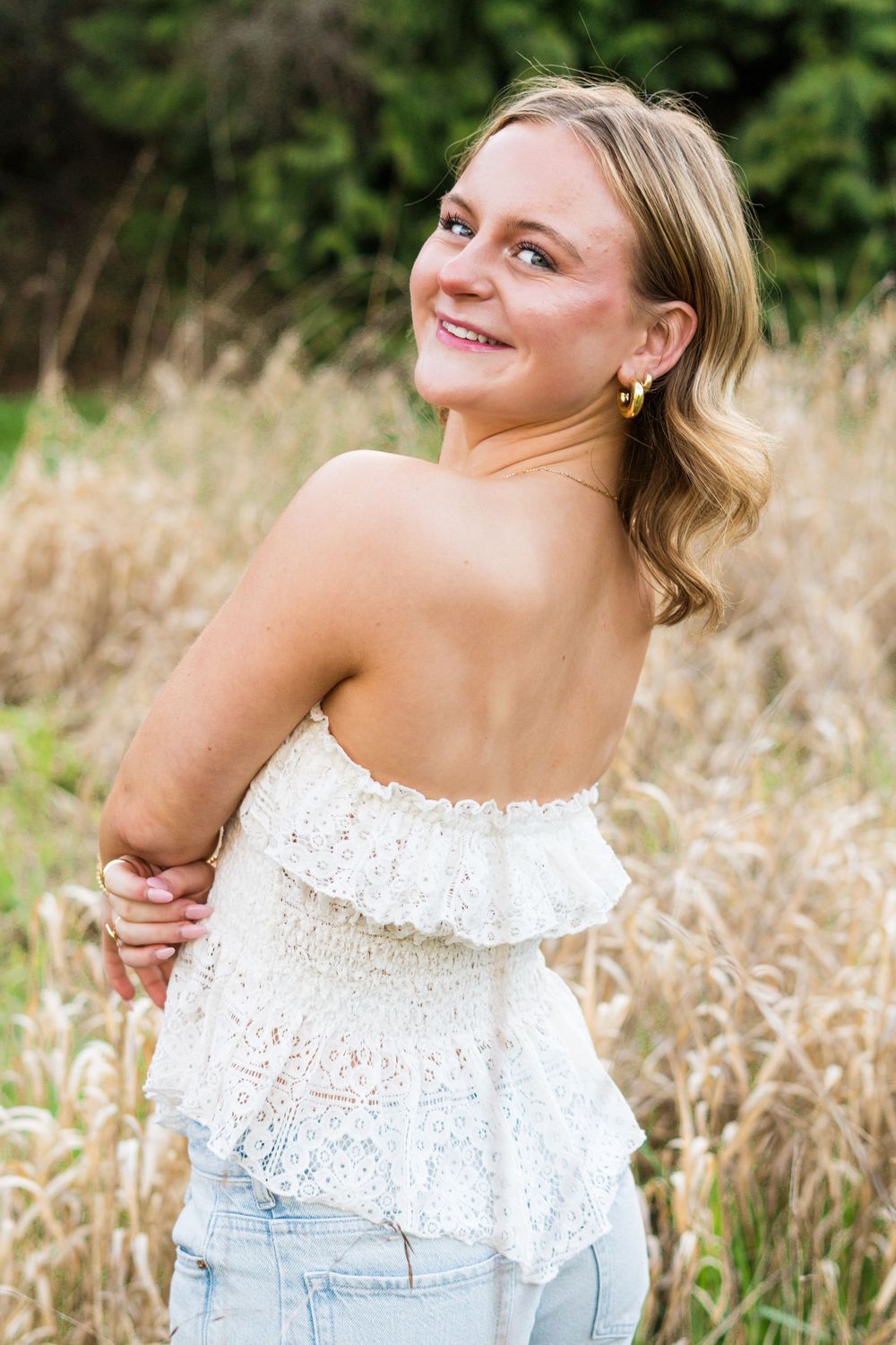 A person in a white eyelet lace top poses joyfully in a natural field setting with golden grass and greenery.