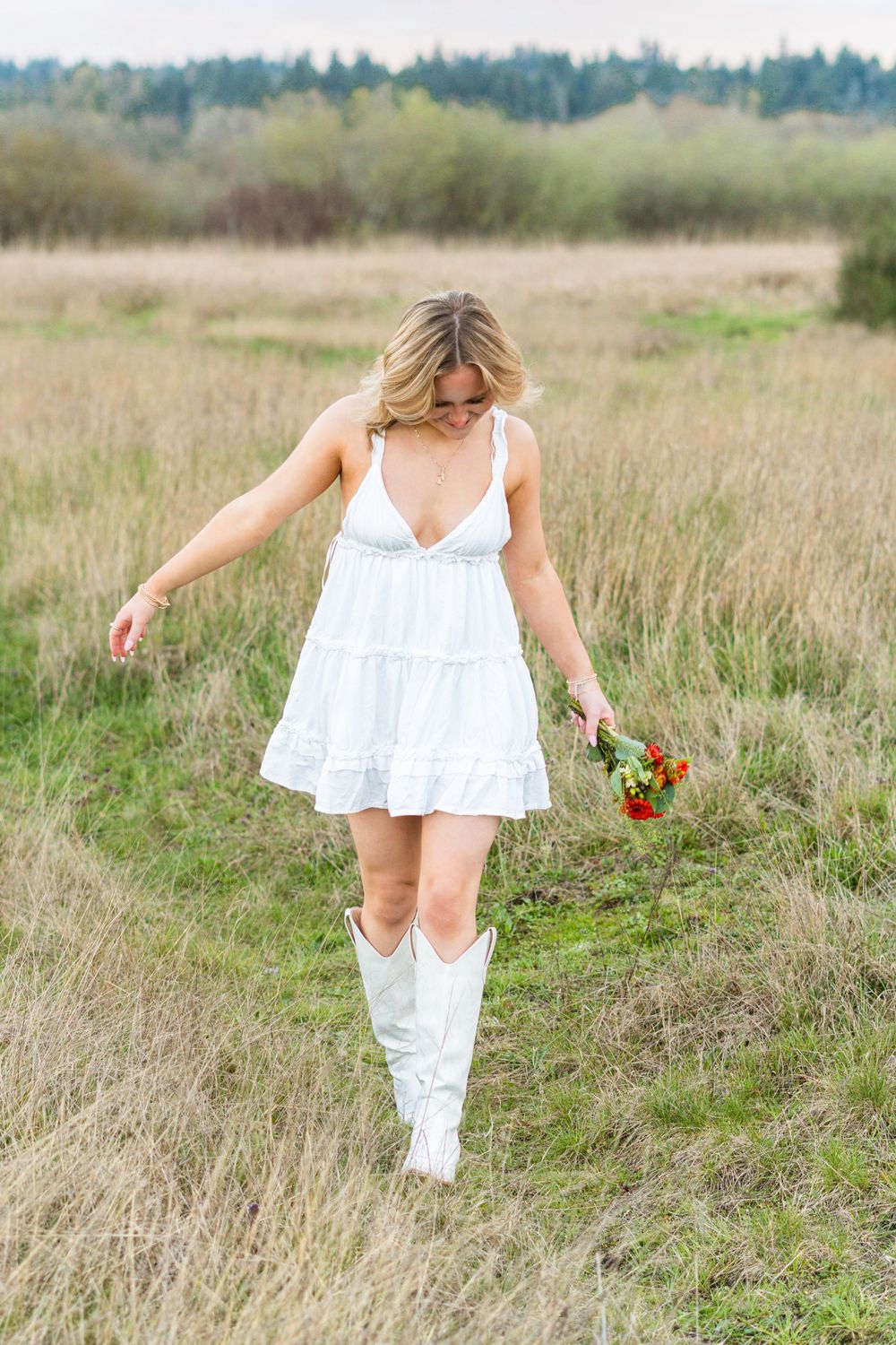 A woman in a white dress walks through a golden field holding red flowers on a sunny day.