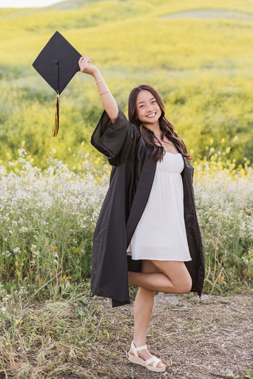 Graduate in cap and gown poses playfully in a field of yellow wildflowers during sunset graduation photoshoot.