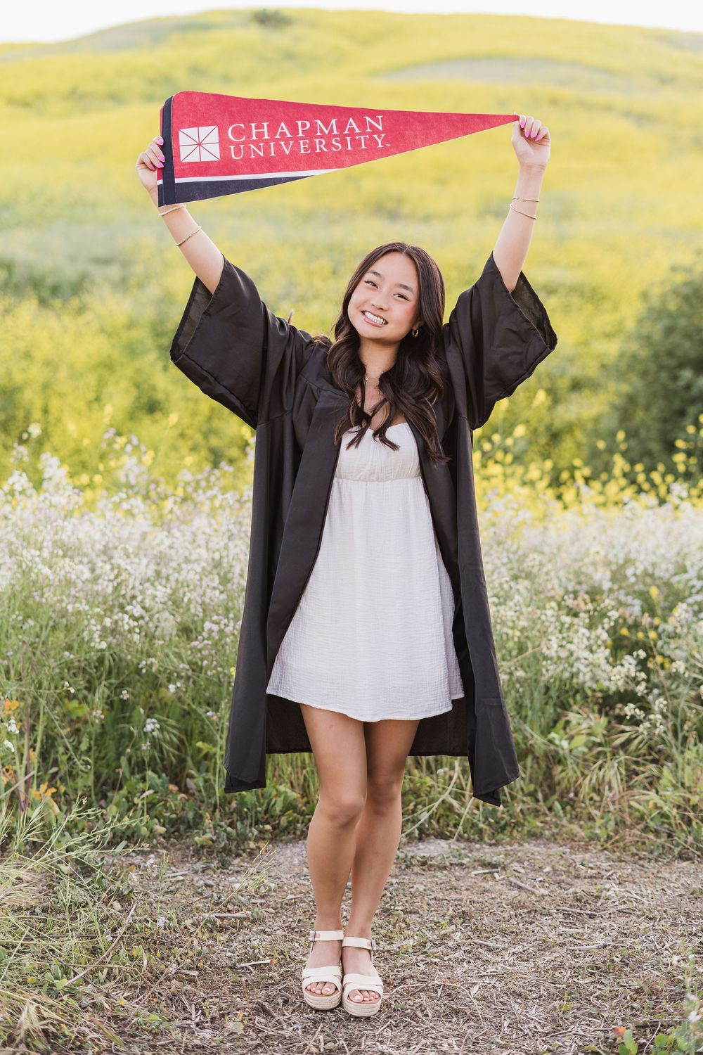 Graduate in cap and gown holding Chapman University banner in a blooming wildflower field.