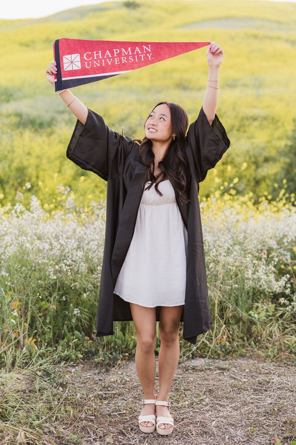 Chapman University graduate celebrates with school pennant among wildflowers during golden hour photoshoot.