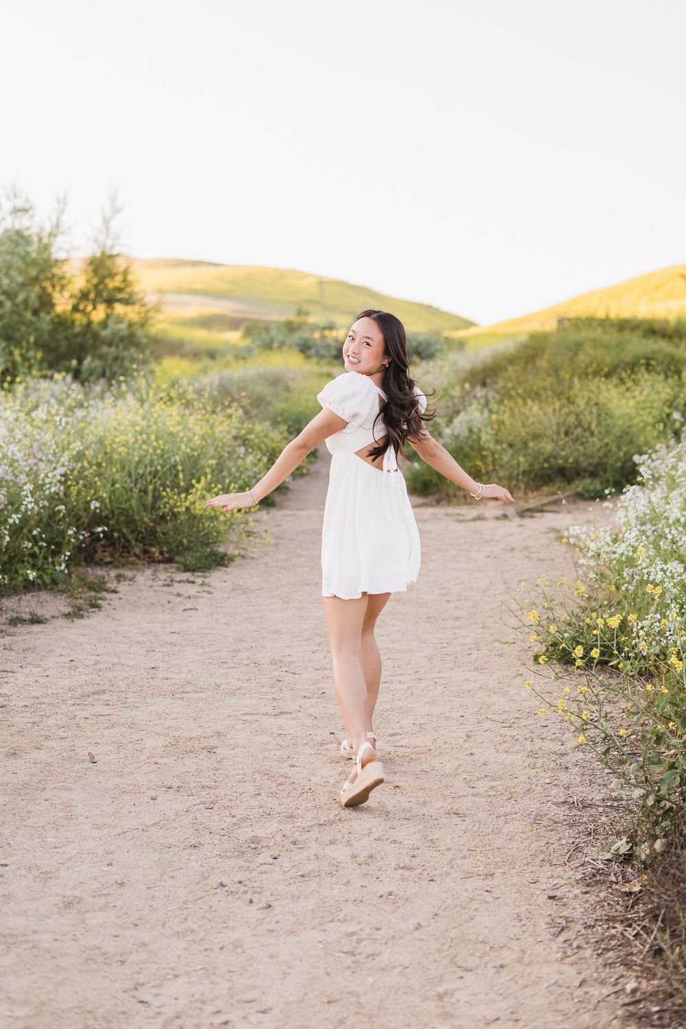Young girl in white dress twirls playfully on a dirt path surrounded by wildflowers at sunset.