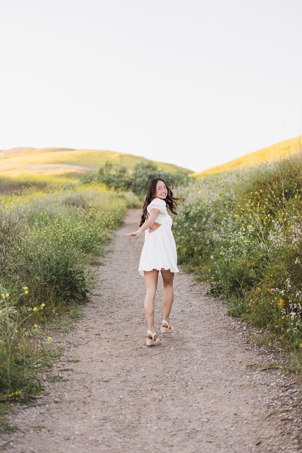 A person in a white dress skips joyfully along a dirt path through a wildflower meadow at sunset.
