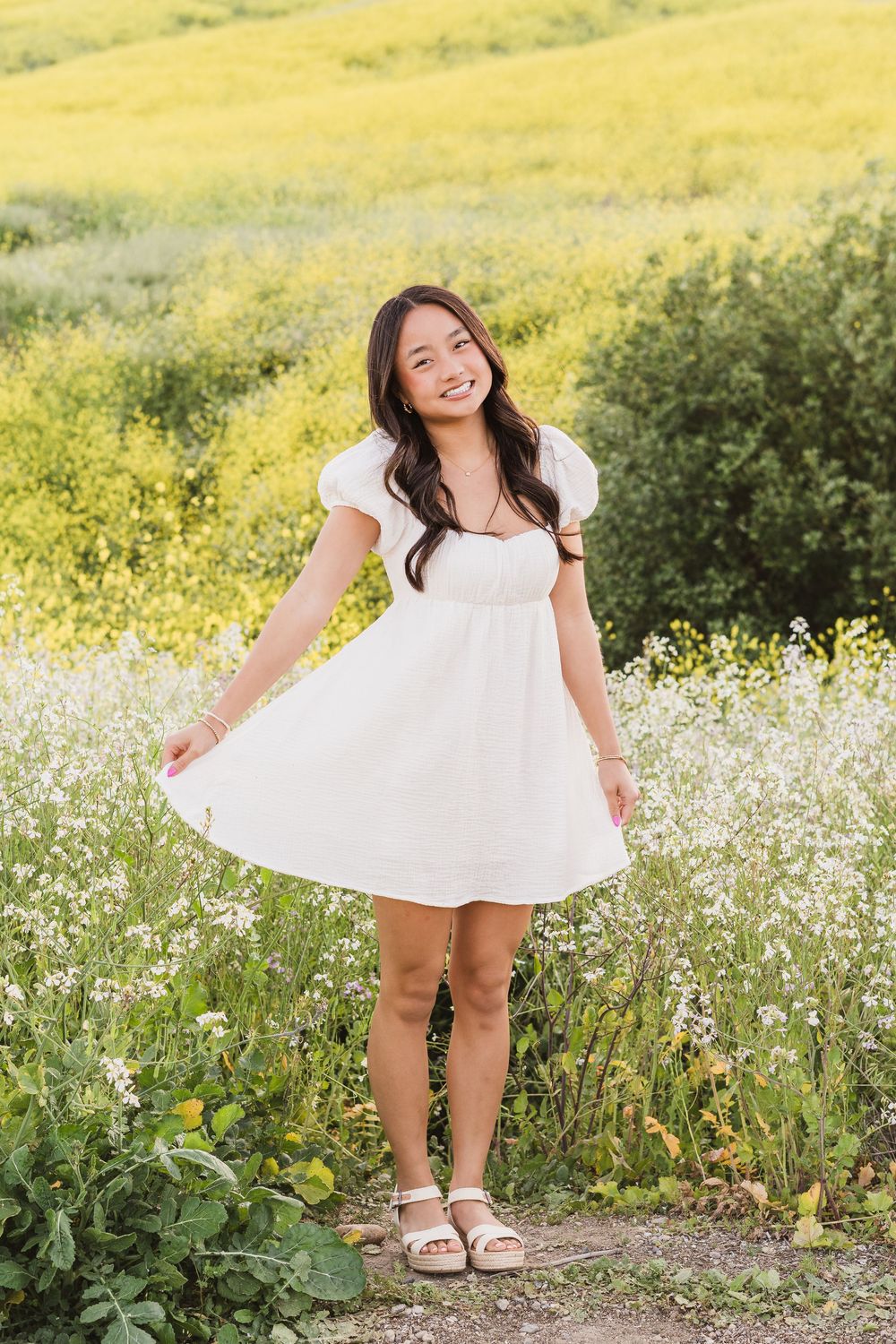 Person in white sundress poses joyfully among yellow wildflowers on a hillside trail.