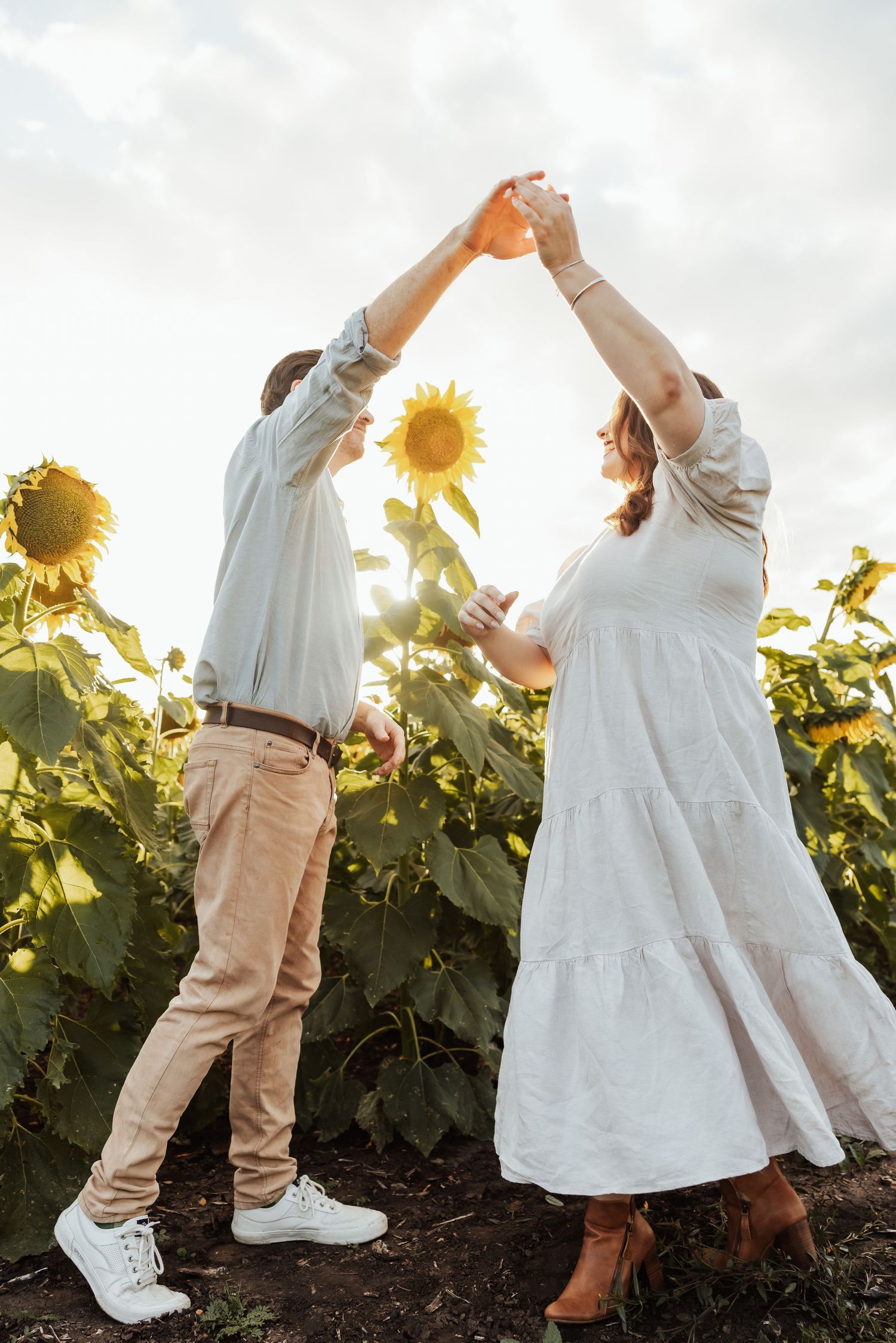 Couple dancing joyfully in a sunflower field at sunset, woman in white dress, man in beige pants.