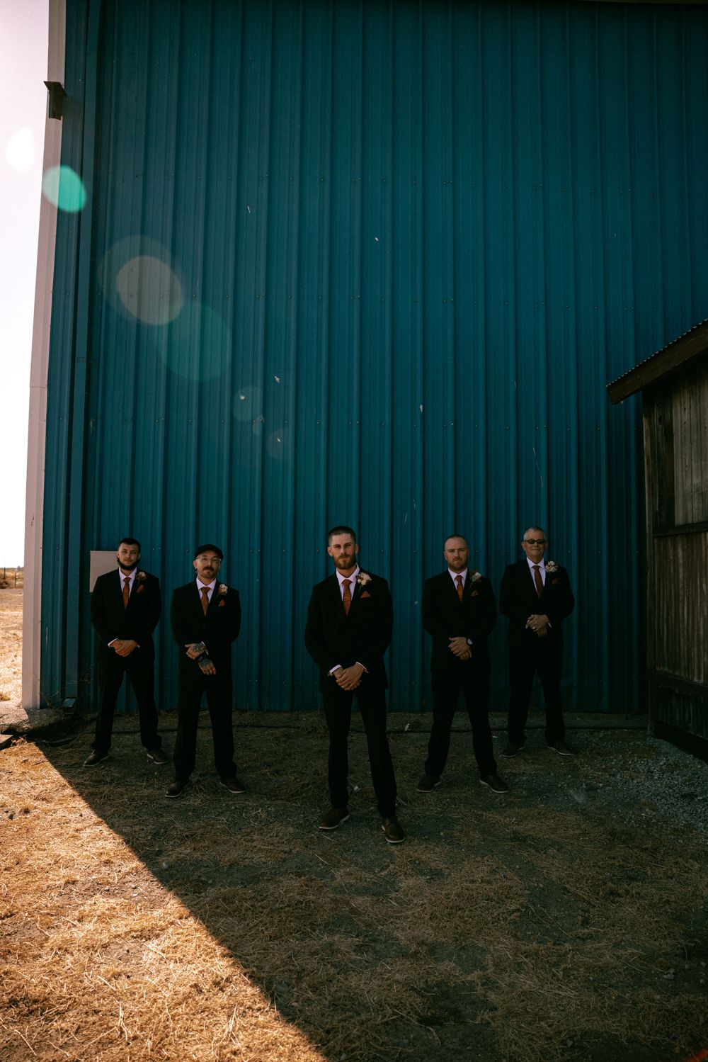 Groomsmen in black suits lined up against a blue wall create a moody artistic wedding photo composition.