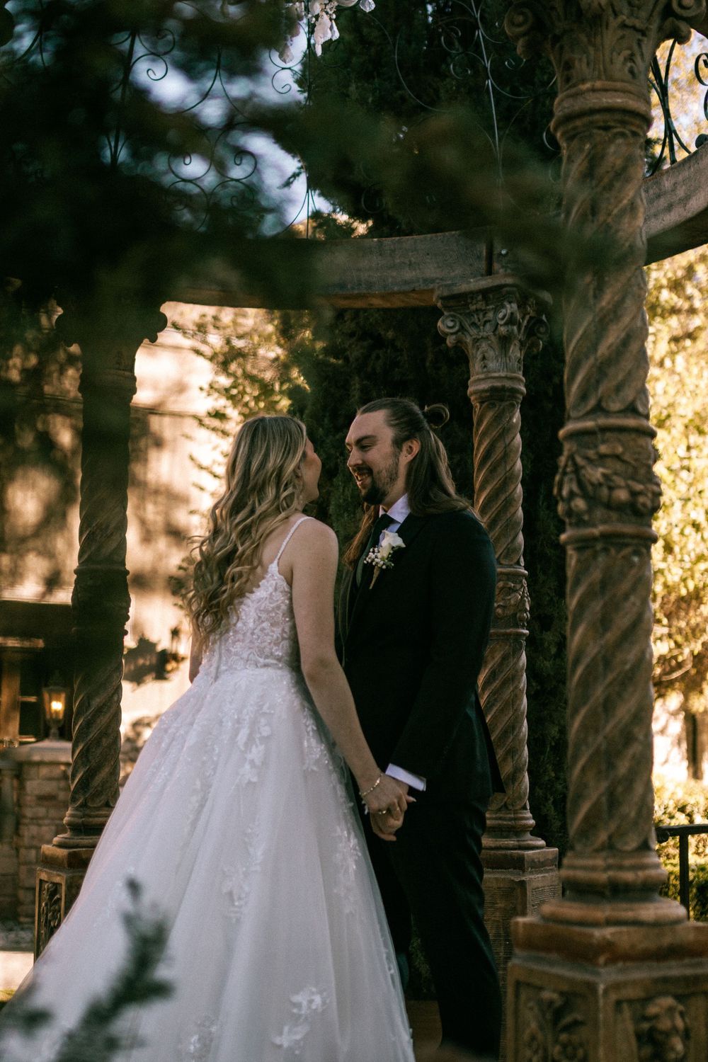A bride and groom share an intimate moment among ornate stone pillars in a sun-dappled forest setting.