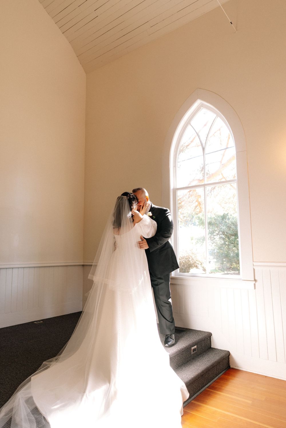 Bride and groom share a romantic moment by an arched chapel window, her flowing veil trailing elegantly on hardwood floors.