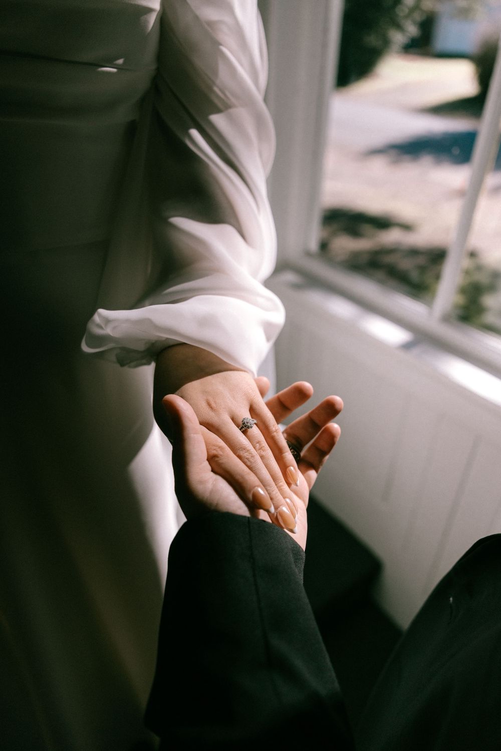 Close-up of a couple holding hands near a window, bride wearing an engagement ring, wedding day moment.