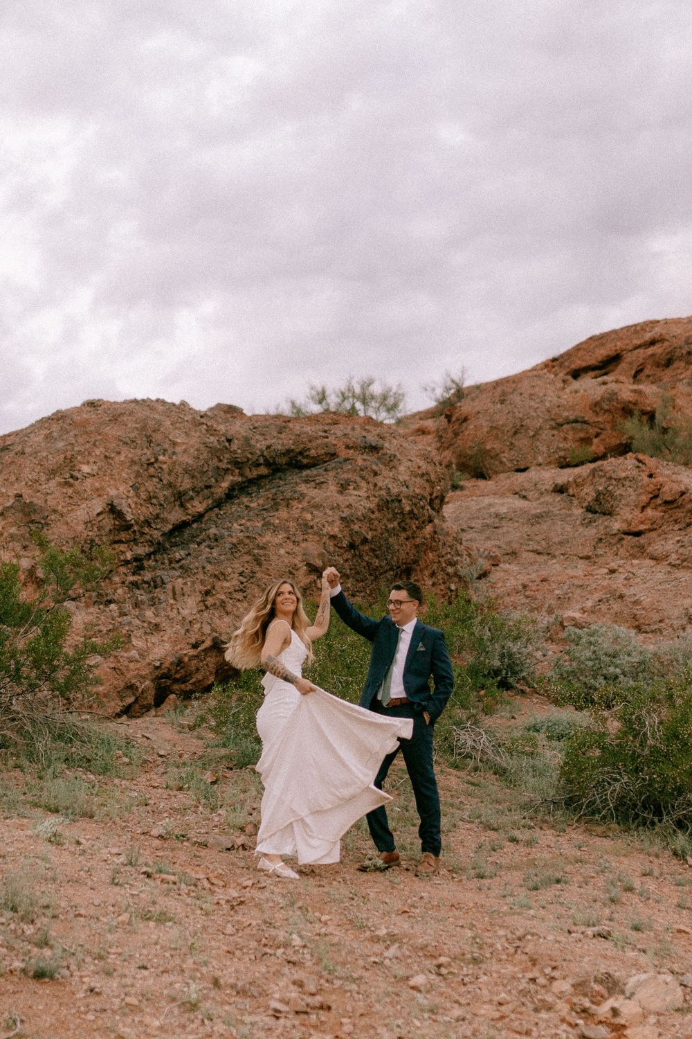 Couple dancing in desert landscape with red rock formations and cloudy sky, bride in flowing white gown, groom in dark suit.