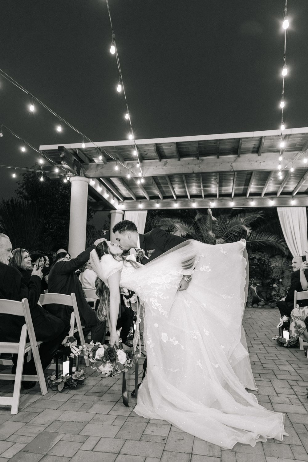Wedding guests gather under string lights on a patio while a bride's white dress flows dramatically in the breeze.