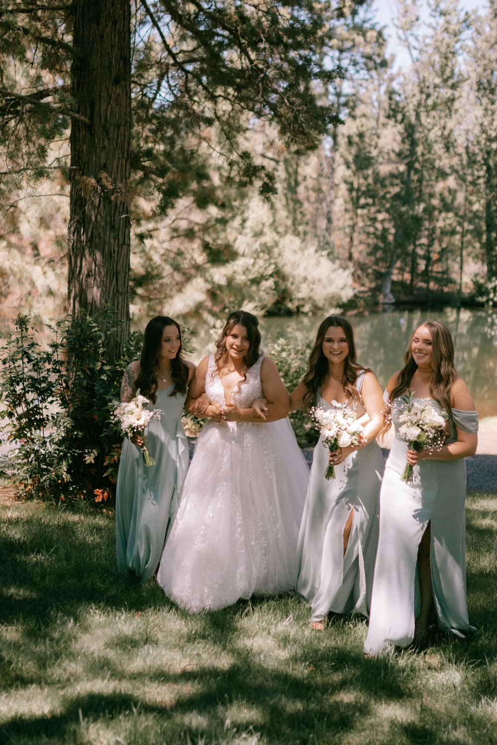 Bridesmaids in soft blue dresses pose with the bride in a white gown against a wooded outdoor backdrop.