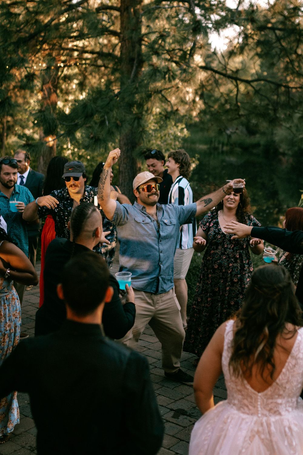 A lively outdoor wedding reception shows people dancing and celebrating on a stone patio surrounded by trees at sunset.