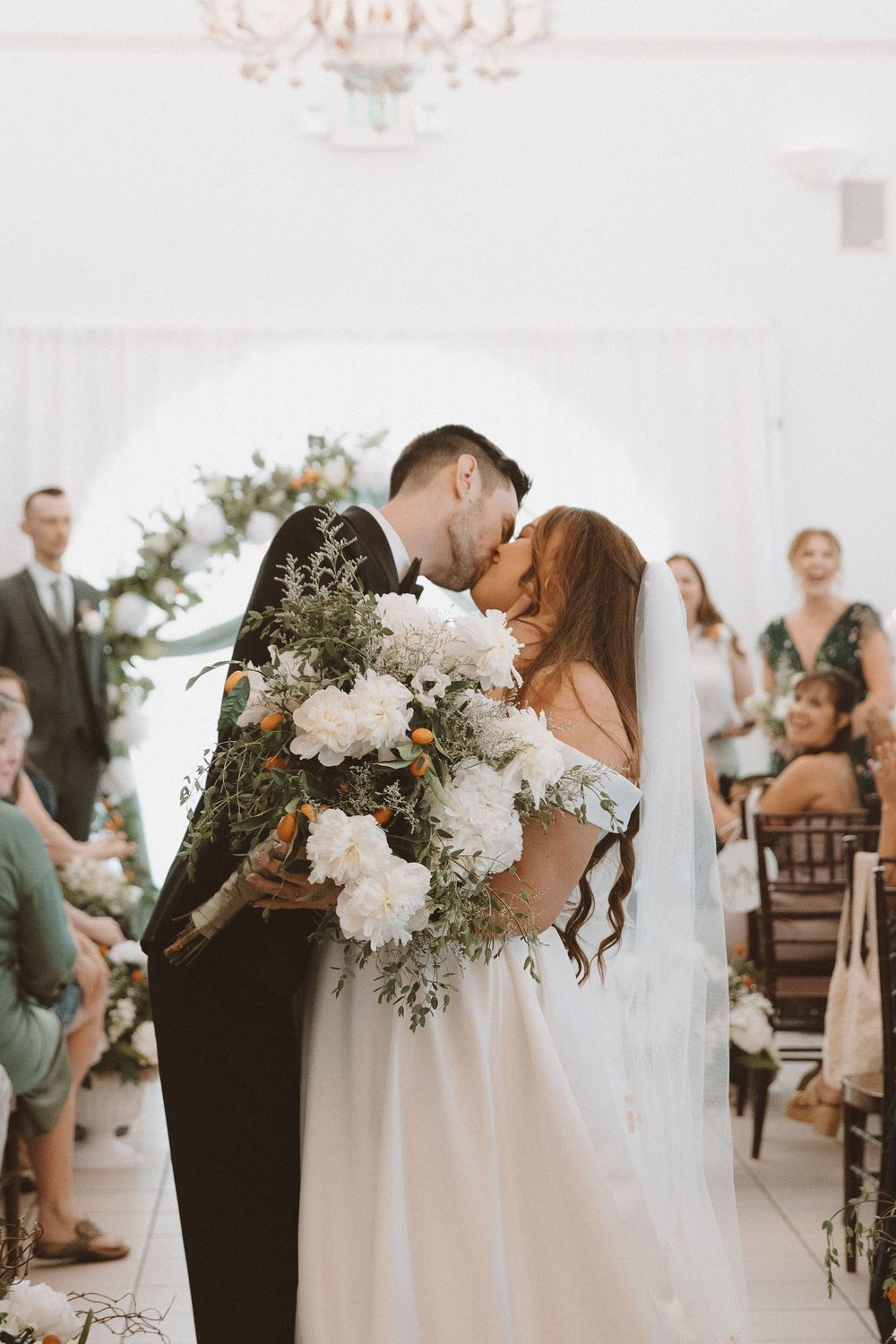 Wedding ceremony kiss with white floral arrangements and guests in attendance.