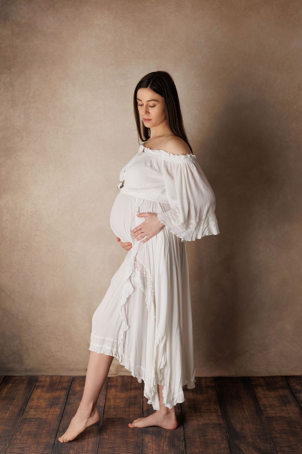 Pregnant woman in white off-shoulder dress cradling baby bump, standing barefoot on wooden floor against beige backdrop.