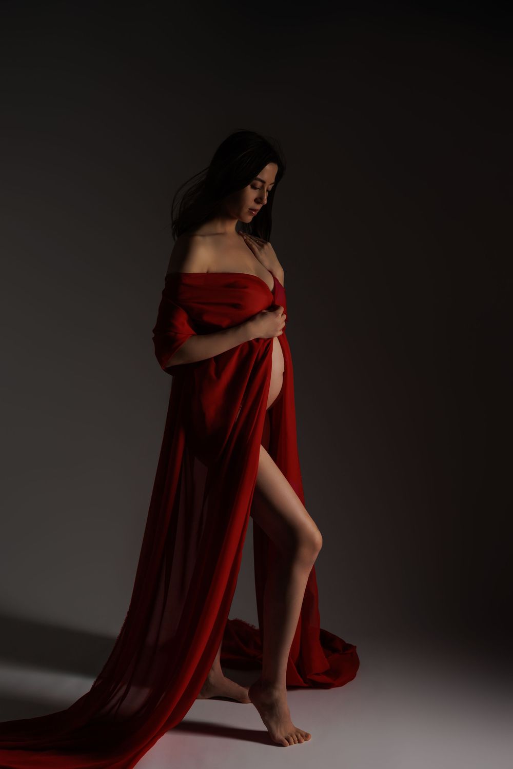 Pregnant woman draped in flowing red fabric poses gracefully in a dark studio setting.