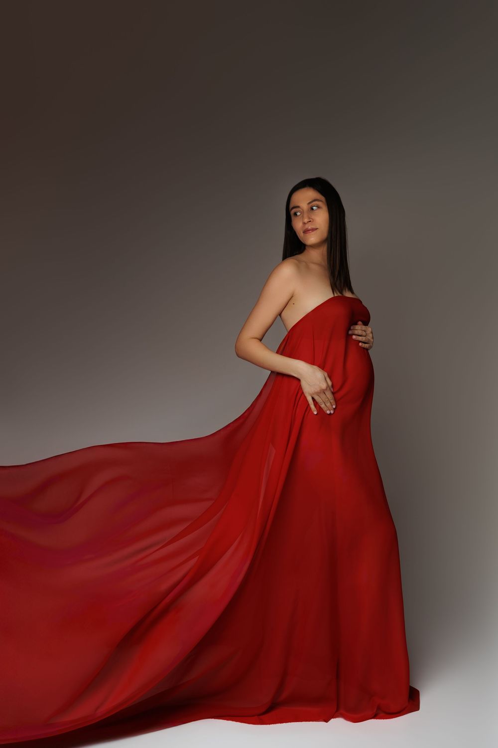 Pregnant woman in flowing red chiffon gown posing gracefully in a studio setting.