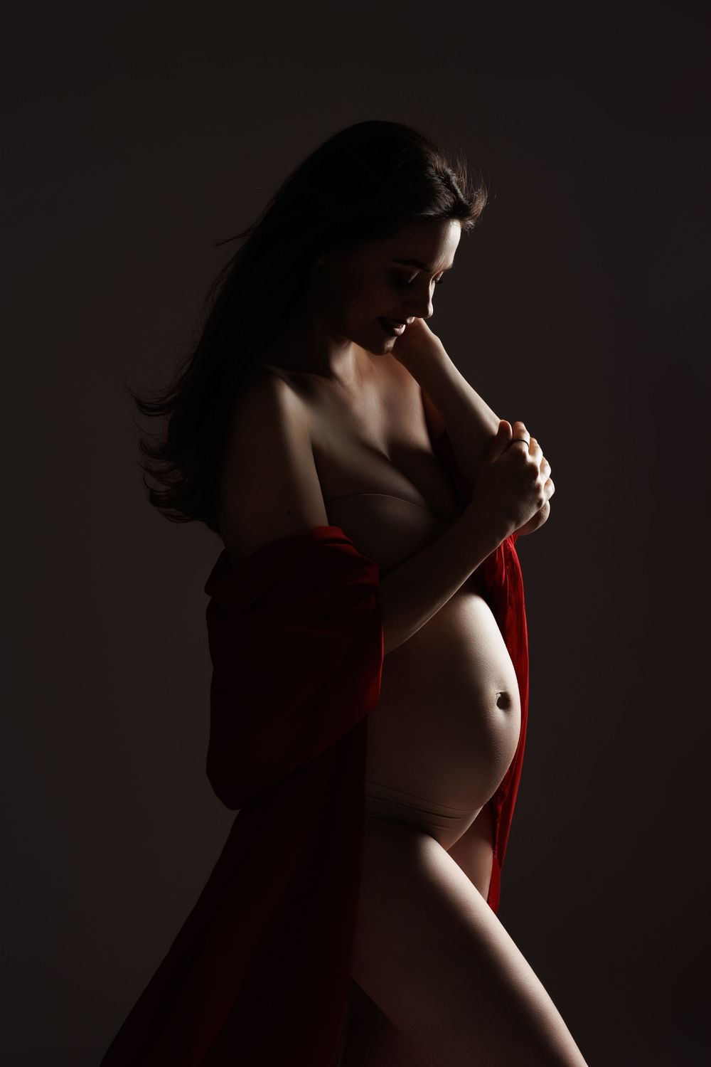Pregnant woman draped in red fabric posing gracefully against dark background in artistic maternity photography.