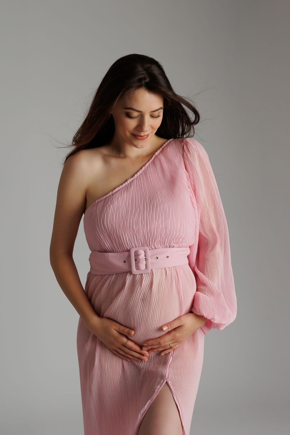 Pregnant woman in pink one-shoulder dress with belt, cradling baby bump, smiling softly against gray background.