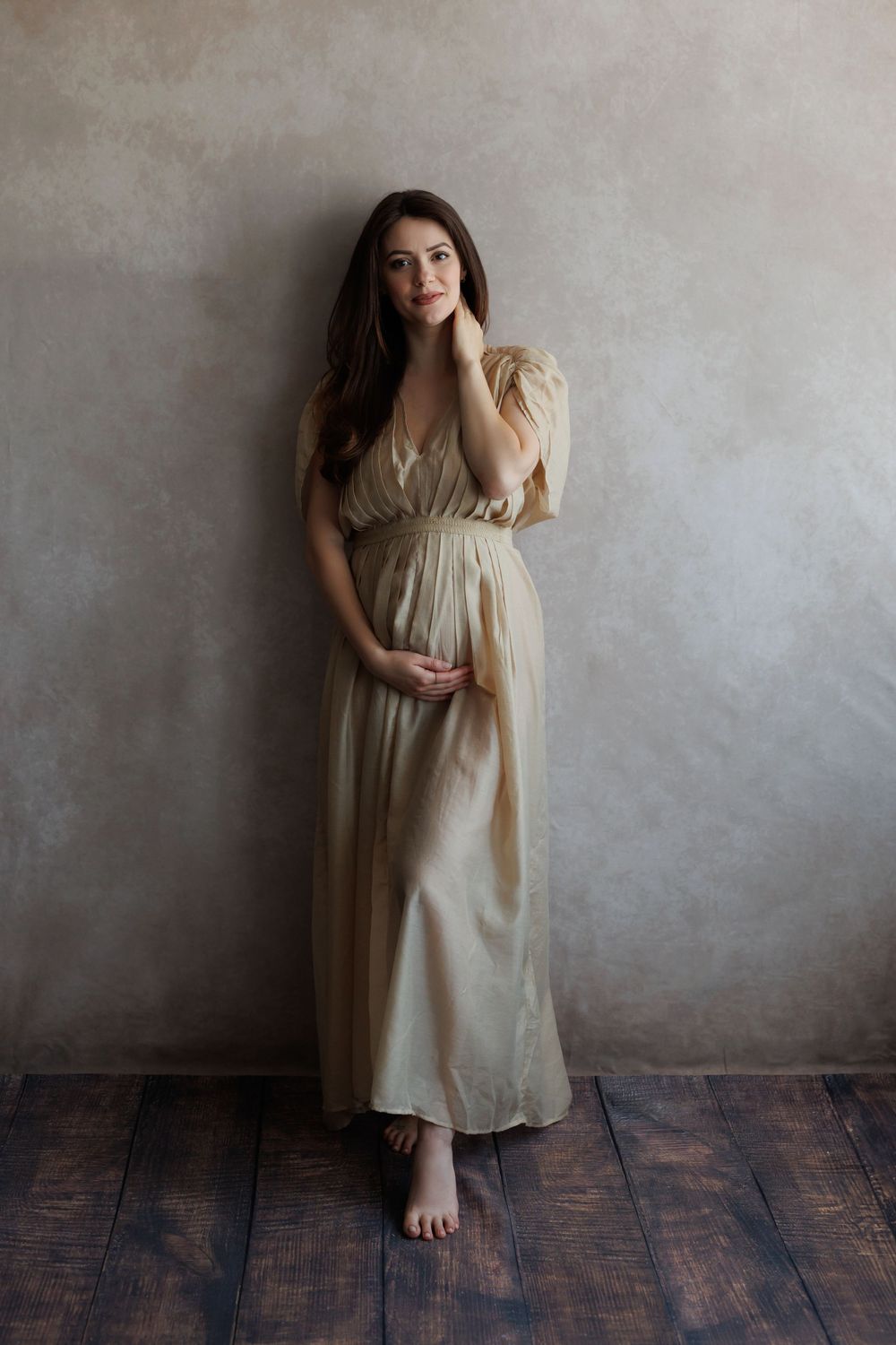 Pregnant woman in elegant beige maxi dress posing barefoot against gray studio backdrop on dark wood floor.