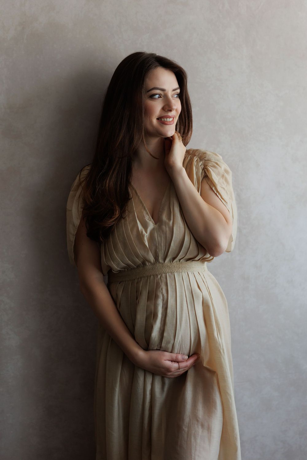 Pregnant woman in elegant golden dress cradling baby bump, smiling softly in a studio portrait.