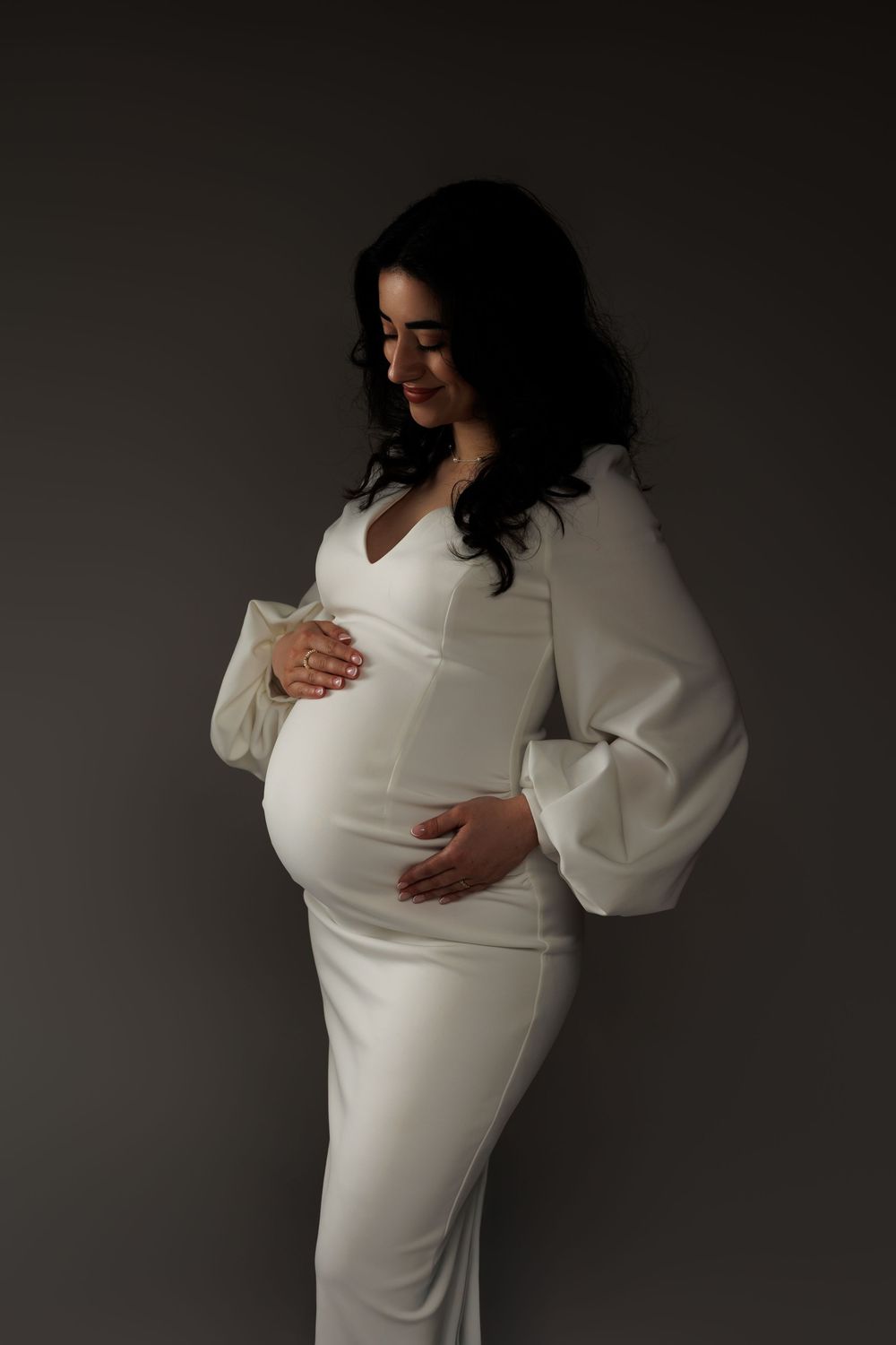 A maternity photoshoot featuring an elegant white dress with bell sleeves against a dark gray backdrop.