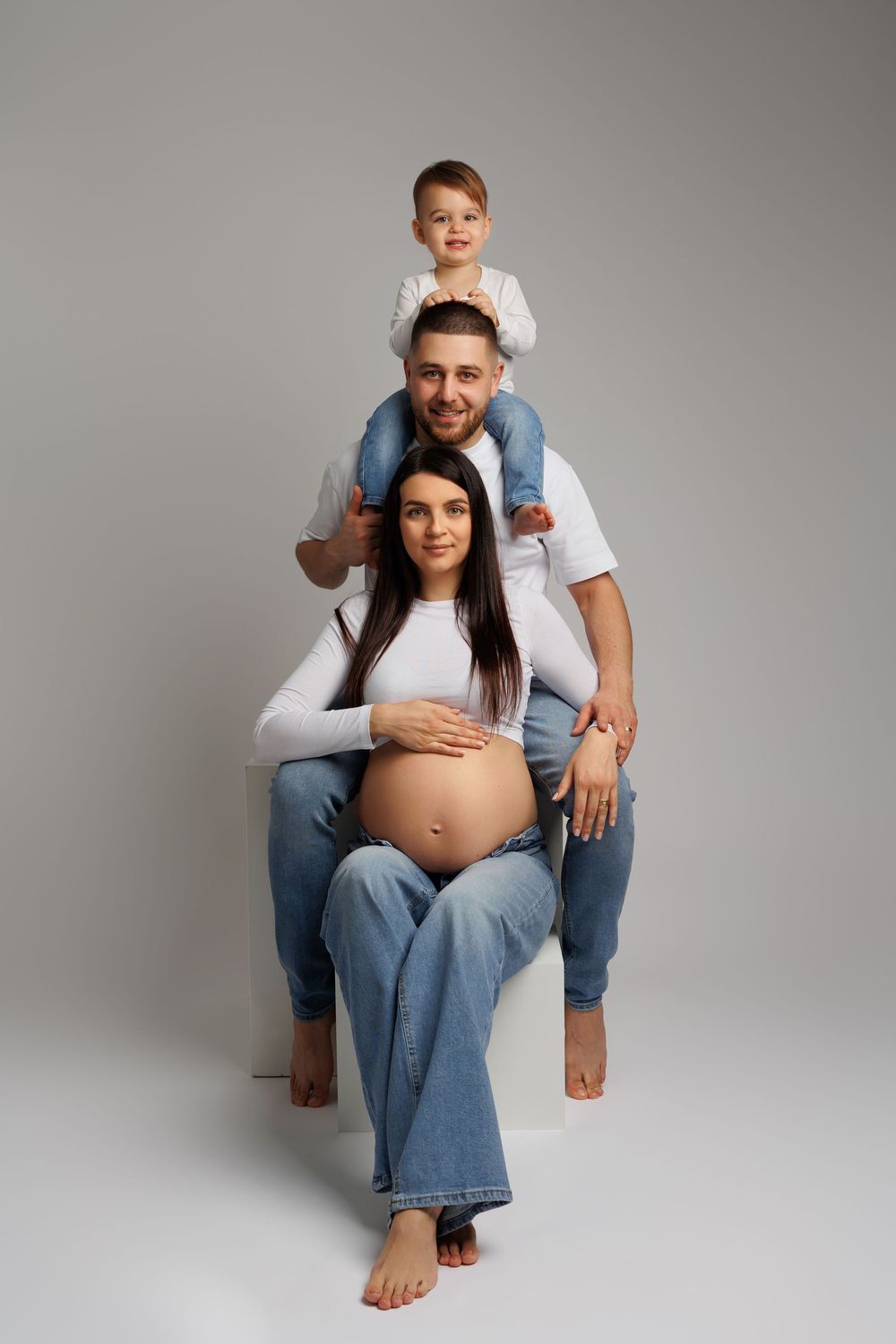 Happy family portrait with pregnant mother, father, and young child on shoulders posing in matching white tops and jeans.