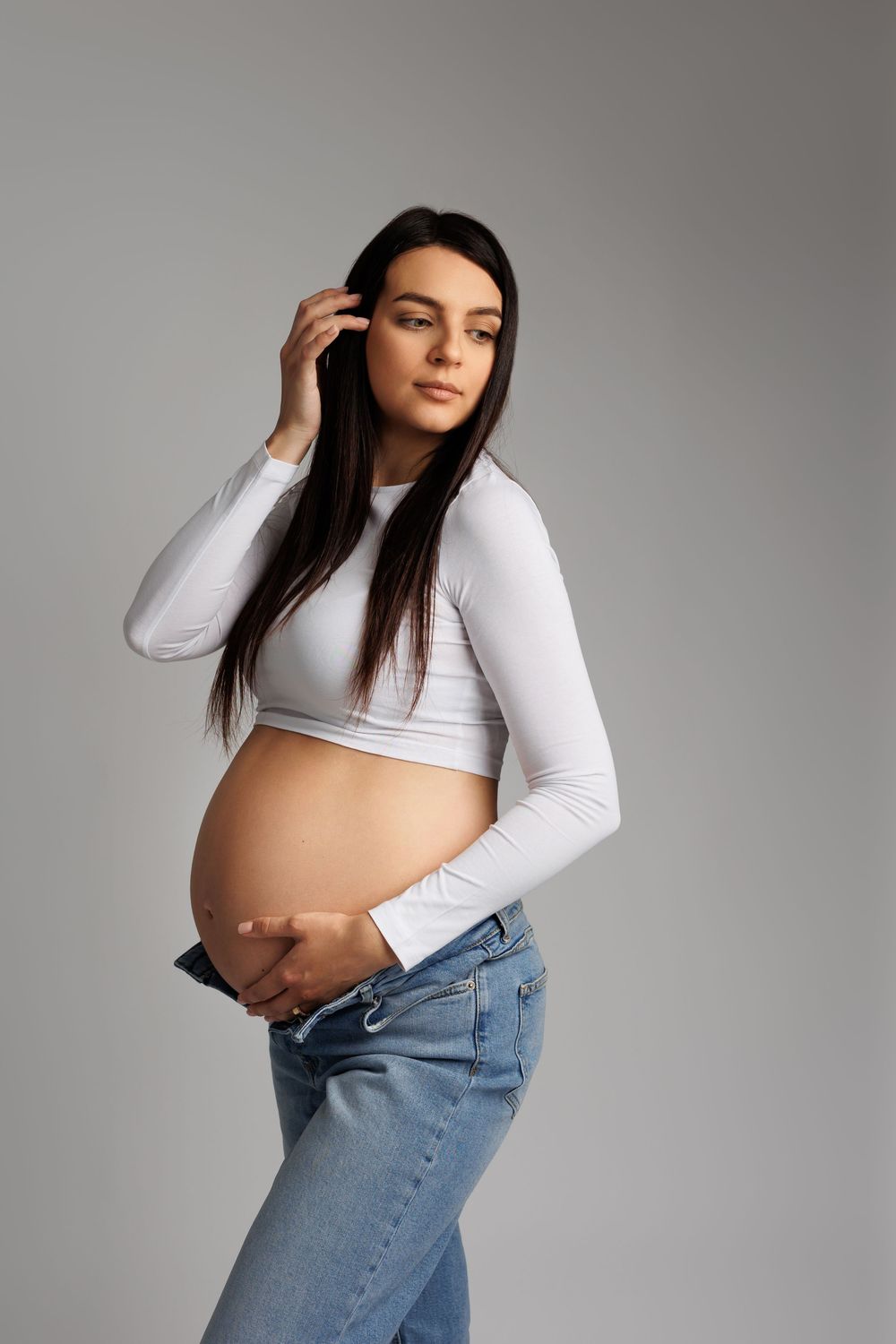 Pregnant woman in white crop top and jeans cradling baby bump, posing against gray background.