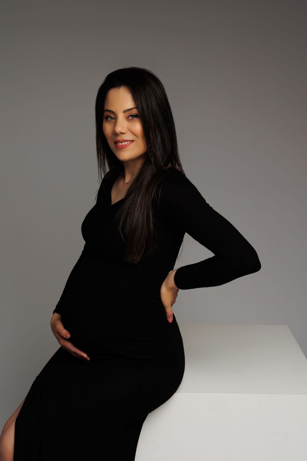 A model in a long-sleeved black dress poses against a gray background in a minimalist studio setting.