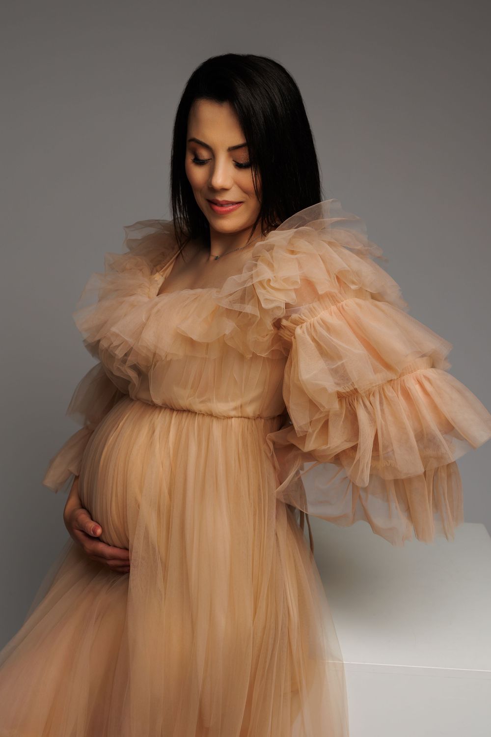 Elegant maternity portrait featuring a flowy peach tulle gown with dramatic ruffled sleeves against a gray backdrop.