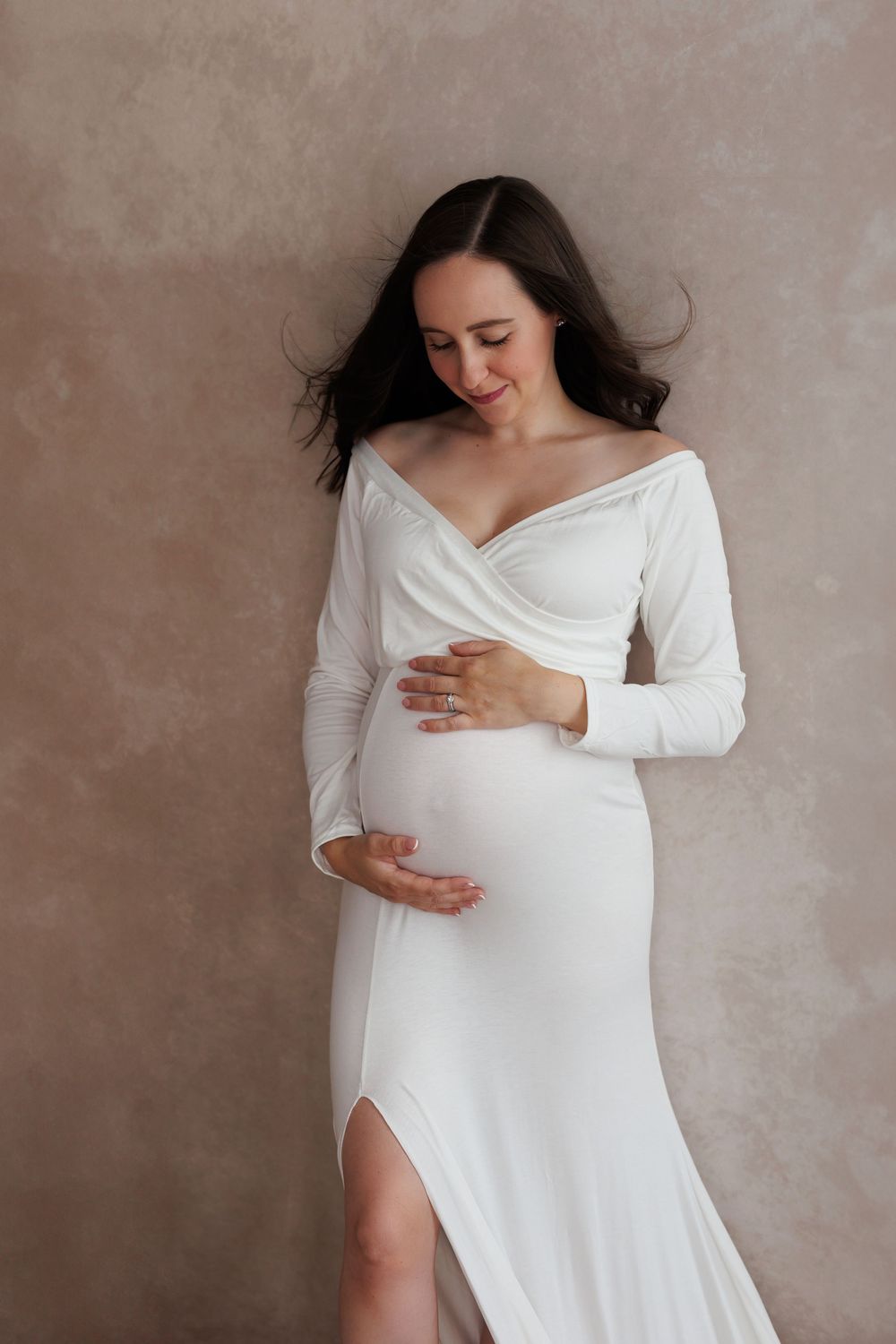 Pregnant woman in elegant white off-shoulder gown cradling her baby bump, smiling softly against a neutral backdrop.