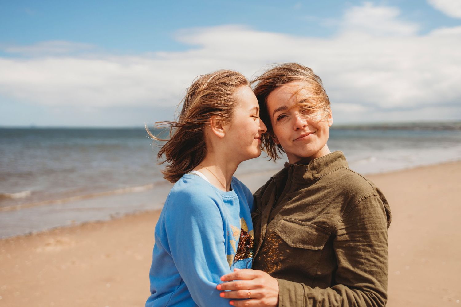 A couple embracing on a windswept beach with blue skies and clouds in the background.