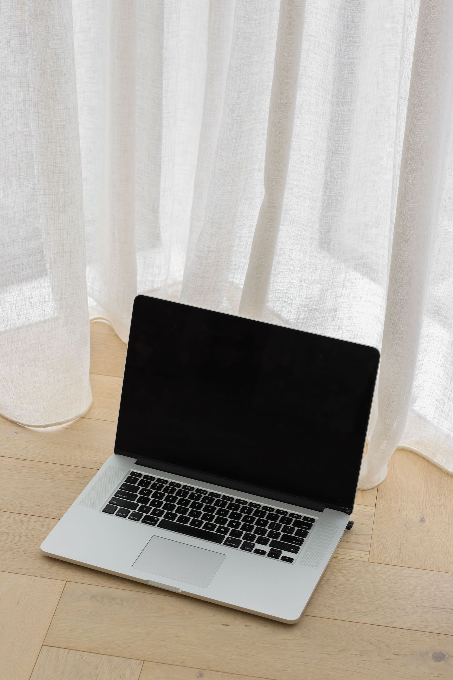 Silver laptop computer positioned against white curtains and wooden floor.
