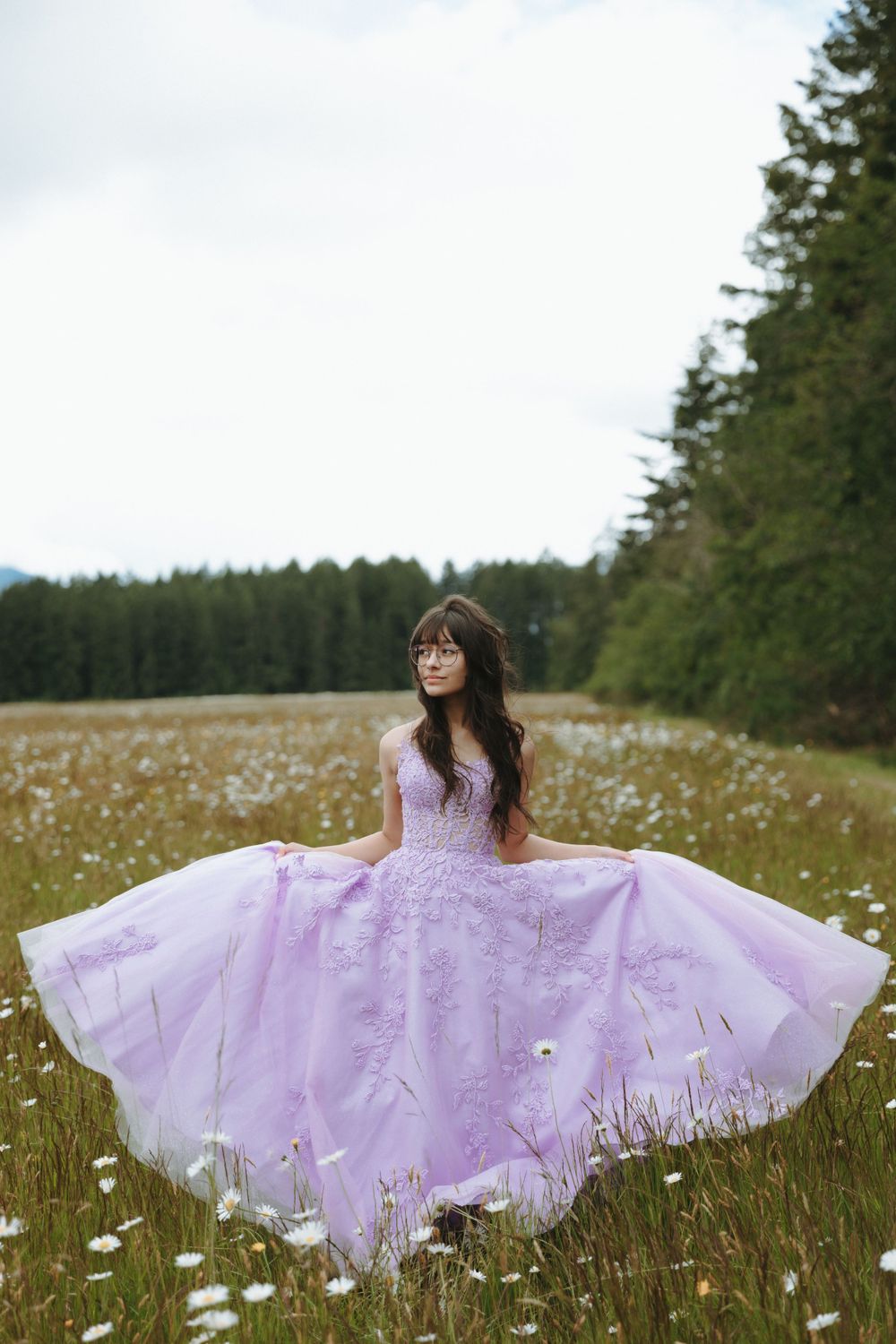 A person in a flowing lilac ballgown twirls elegantly in a wildflower meadow surrounded by evergreen forest.