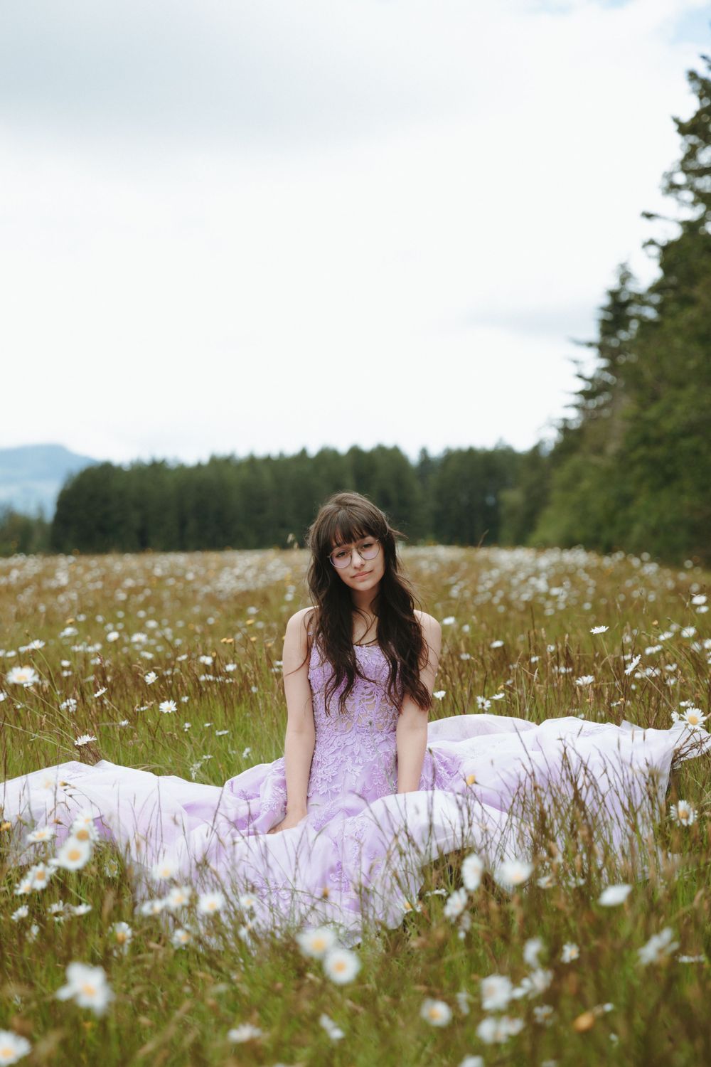 A person in a pink dress sits in a mountain meadow filled with white wildflowers on a cloudy summer day.